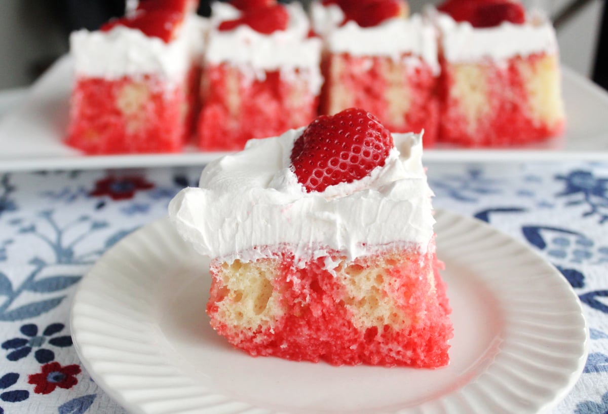 A serving of strawberry poke cake on a white plate with a try of poke cake portions in the background.