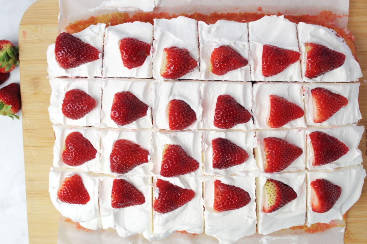 A strawberry poke cake cut into 24 pieces with a strawberry on each piece.