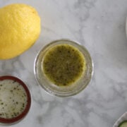 An open jar of three ingredient Greek Salad Dressing. Lemon, olives and sliced tomatoes to the side.