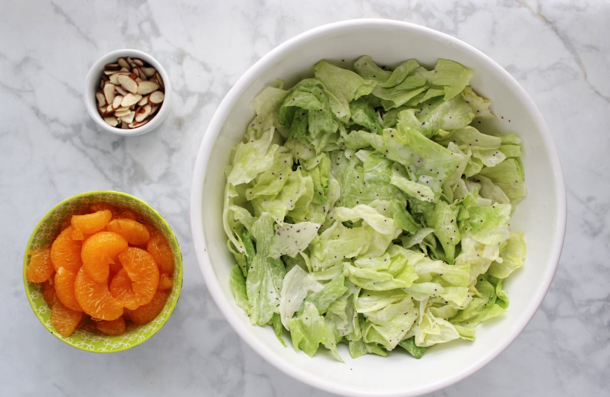 Lettuce lightly covered in poppyseed dressing in a large white bowl.  Mandarin oranges and sliced almonds in small containers to the side of the large bowl.