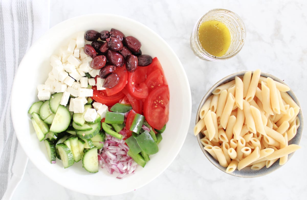 Large bowl with cut up tomatoes, cucumbers, green peppers, red onions, feta cheese and olives. Smaller bowl including cooked pasta. Mason jar with homemade Greek Dressing.