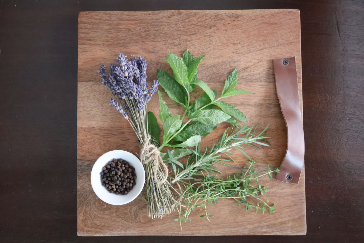 a cutting board displaying fresh mint, rosemarry and thyme plus a small container of peppercorns and a bundle of dried lavender.