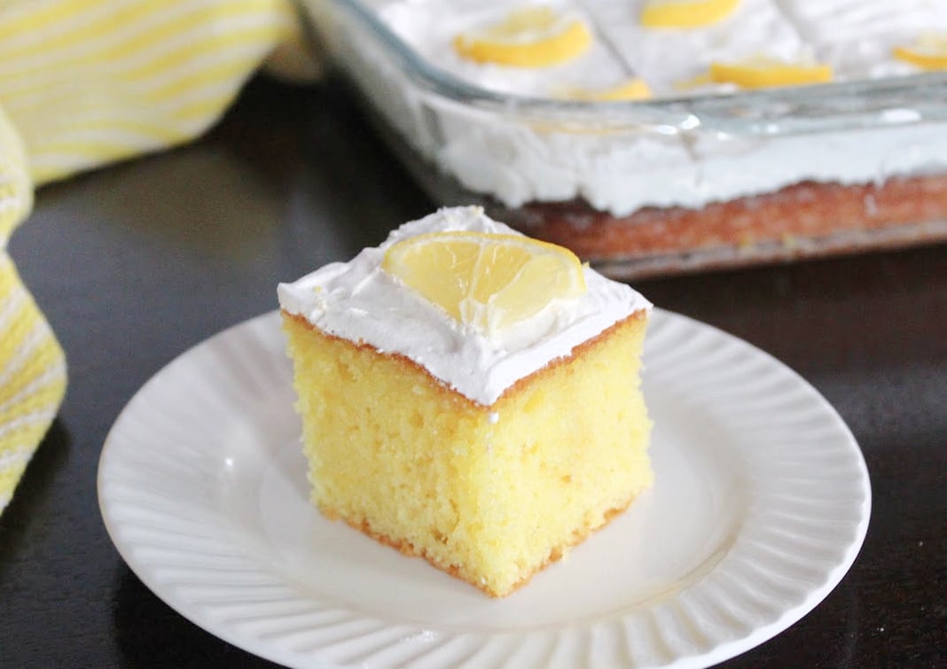 Lemony lemon poke cake on a white plate.  Poke cake is garnixhed with whiped topping and a ¼ slice of lemon.