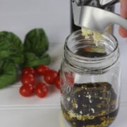 garlic being pressed into a jar with oil and balsamic vinegar to make balsamic vinaigrette dressing. Tomatoes and fresh basil leaves are in the background.