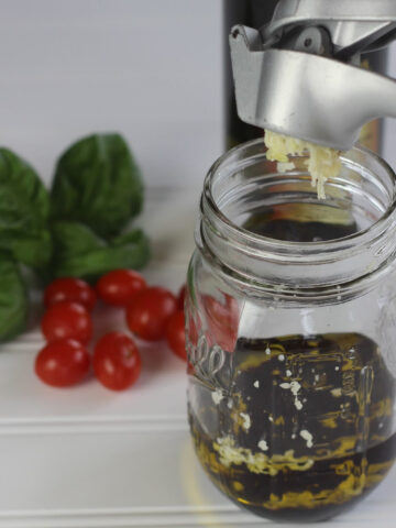 garlic being pressed into a jar with oil and balsamic vinegar to make balsamic vinaigrette dressing. Tomatoes and fresh basil leaves are in the background.