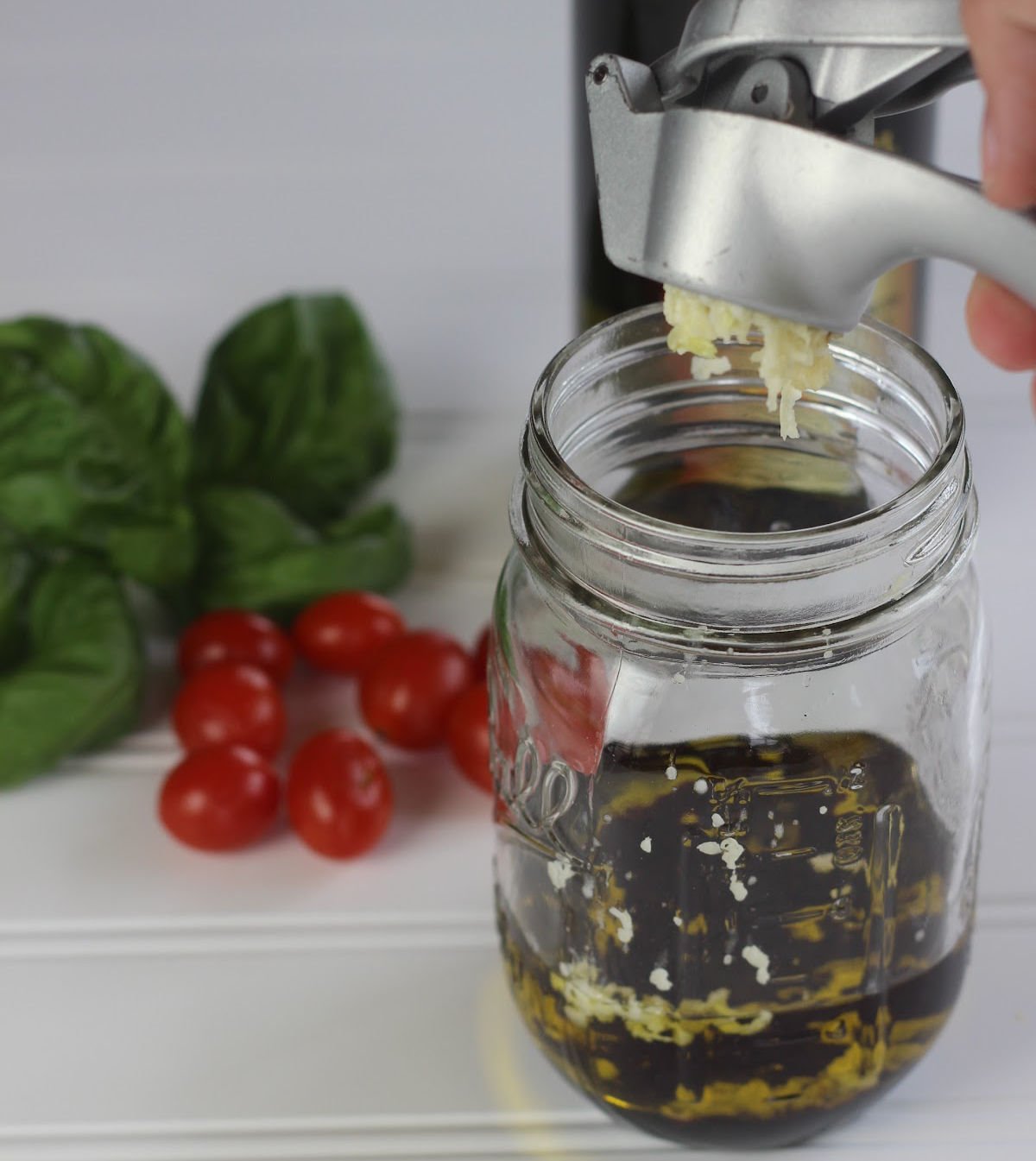 garlic being pressed into a jar with oil and balsamic vinegar to make balsamic vinaigrette dressing.  Tomatoes and fresh basil leaves are in the background.