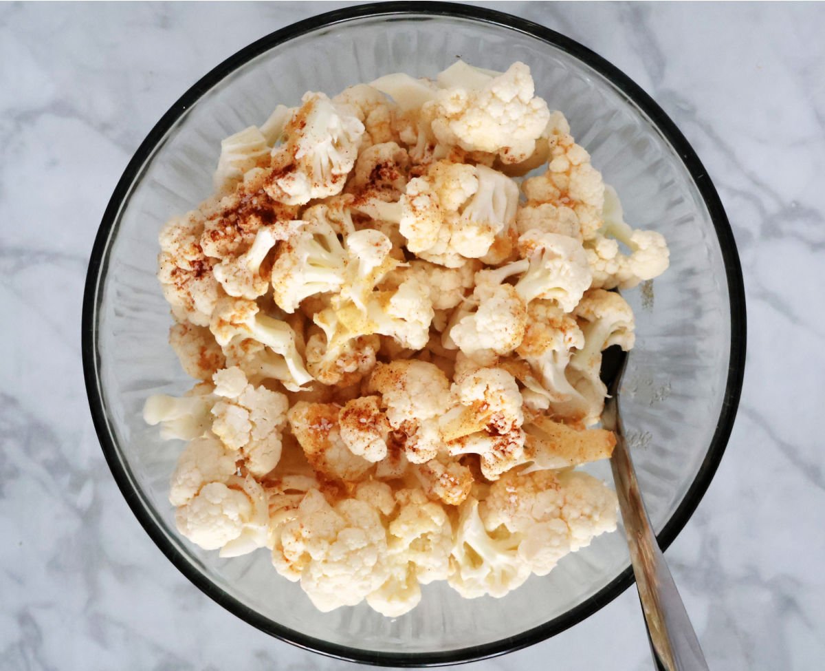 A large clear bowl with cauliflower florets that are visibly seasoned.