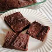 three pieces of pumpkin brownies on a white plate with a baking dish full of two ingredient brownies in the background.