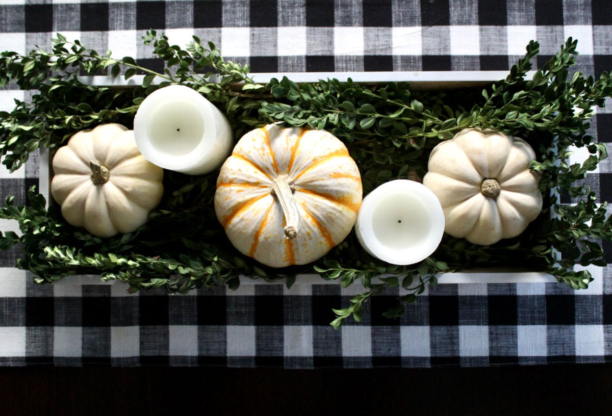A centerpiece in a wooden tray containing white pjmpkins, white flameless candles and boxwood clippings on a black and white buffalo plaid runner