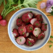 a bowl with roasted radishes. Fresh radishes and flowering chives surround the bowl for color.