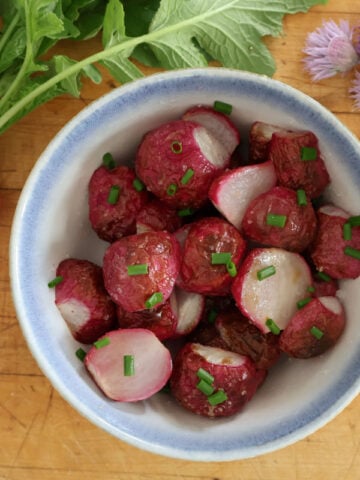 a bowl with roasted radishes. Fresh radishes and flowering chives surround the bowl for color.