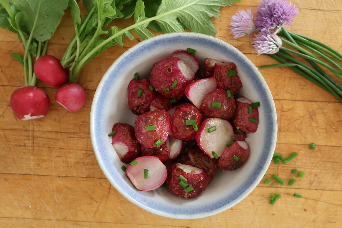 a bowl with roasted radishes. Fresh radishes and flowering chives surround the bowl for color.