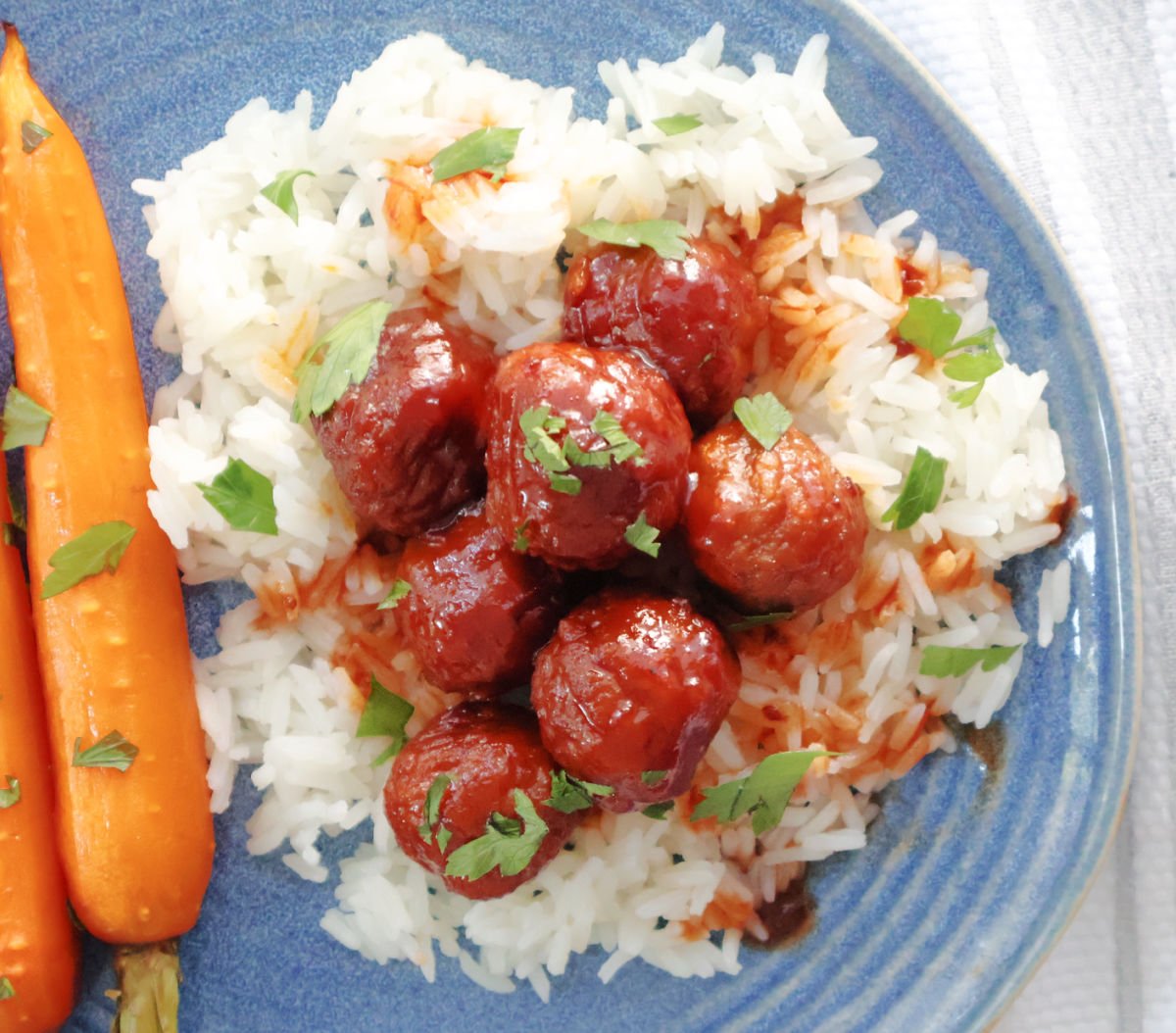 meatballs on top of a bed of rice with a sauce on the meatballs and some chopped parsley as the garnish. Roasted carrots are also on the plate