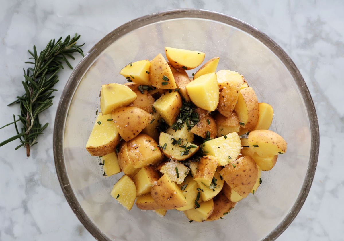 Cut up potatoes with olive oil, minced rosemary and course salt added, ready to be mixed in. Sprig of rosemary sits on the counter beside the bowl.