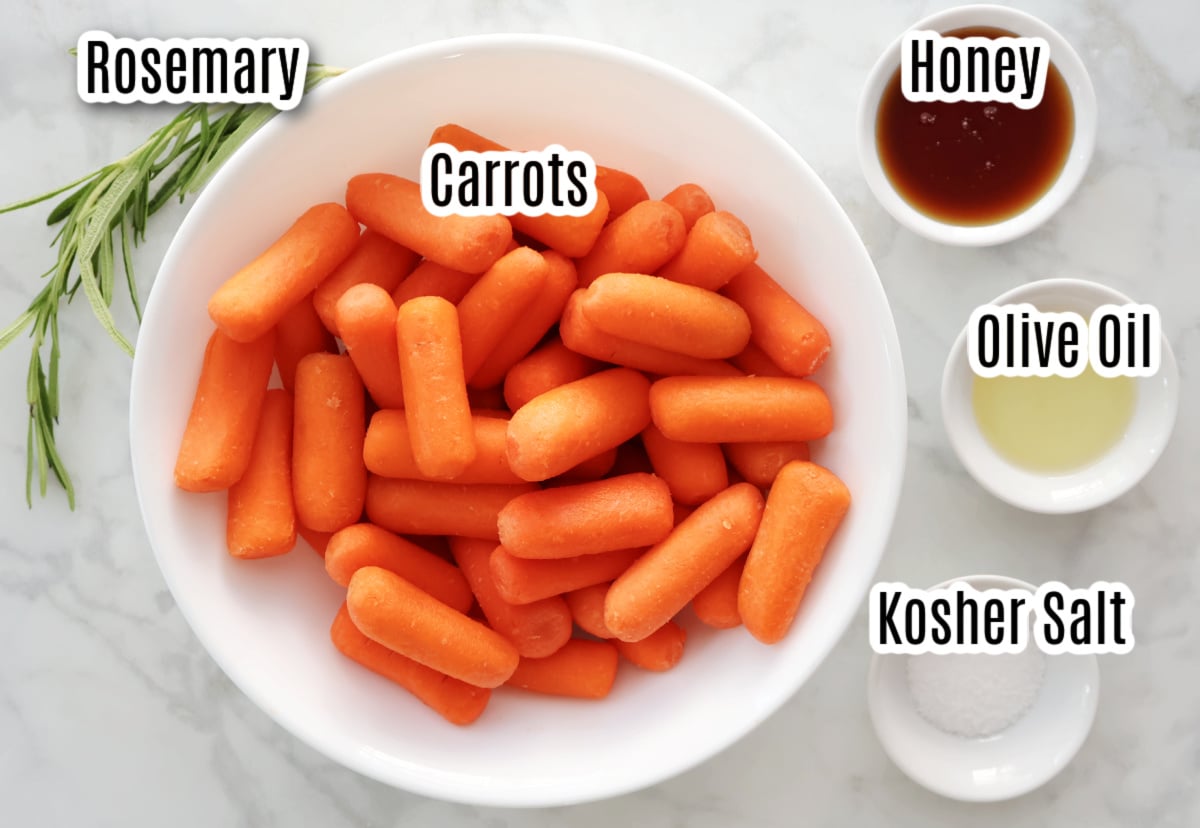 ingredients for baby carrots made in the air fryer.  Includes a bowl of baby carrots, small bowls containing honey, olive oil and kosher salt. a sprig of rosemary is beside the carrots.