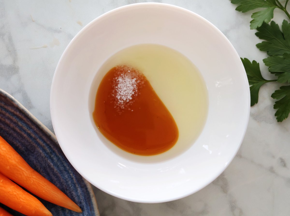 small white bowl with olive oil, honey and Kosher salt ready to be mixed. Bunch of peeled carrots are off to the side.