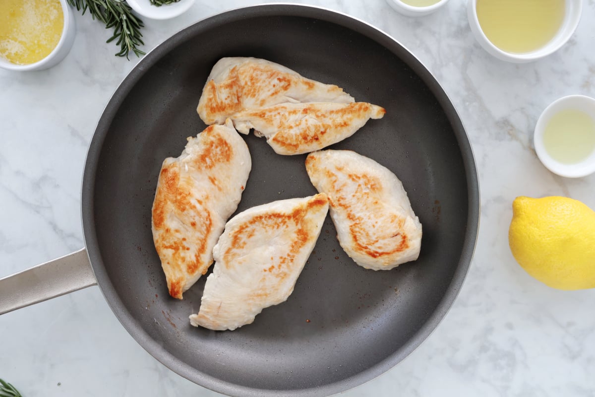 4 pieces of chicken browned in a frying pan. Ingredients for the sauce can be seen and they include lemon, chicken broth, oil, and fresh rosemary.