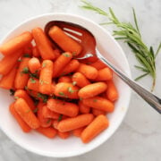 baby carrots with oil, Kosher salt and rosemary mixed together with a large spoon in a white bowl. Sprig of rosemary beside bowl for color