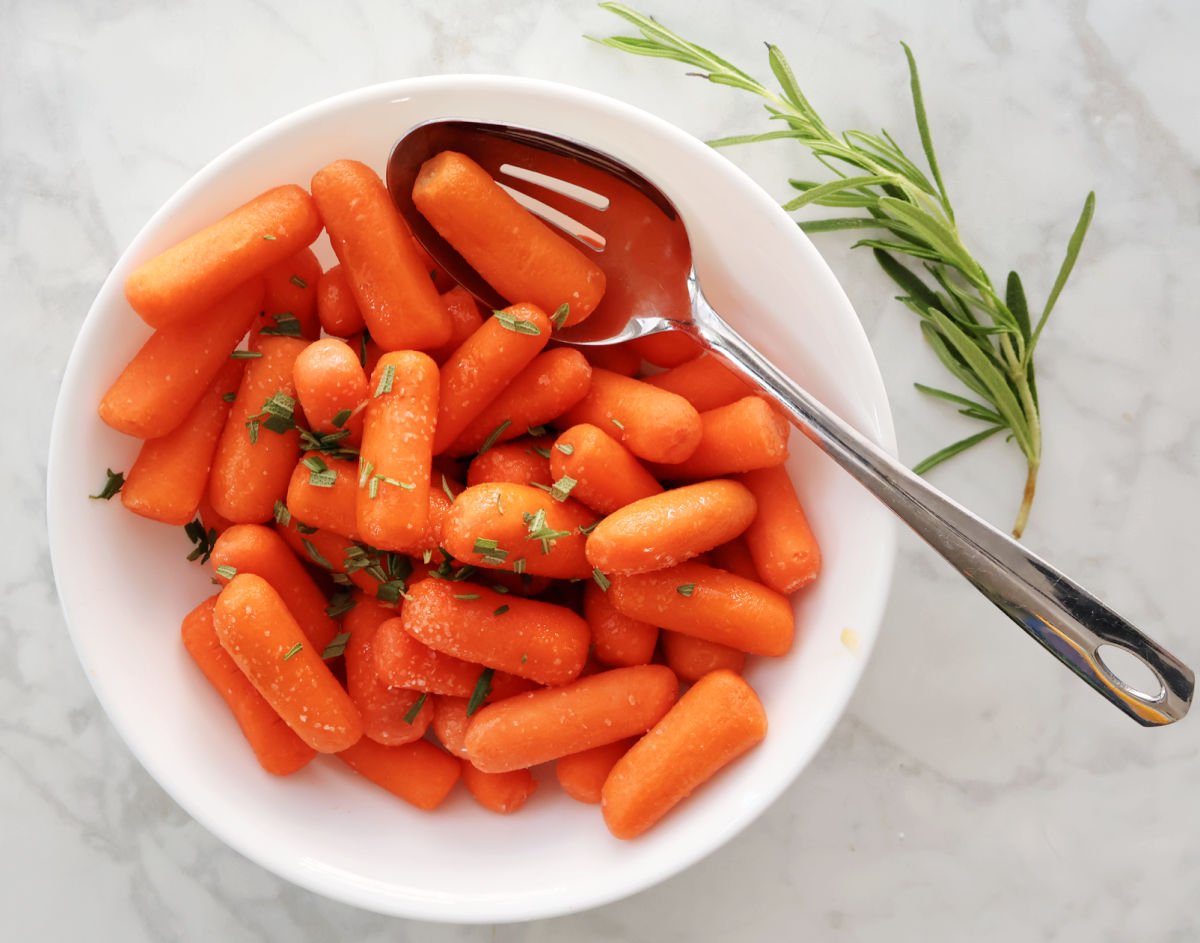 baby carrots with oil, Kosher salt and rosemary mixed together with a large spoon in a white bowl. Sprig of rosemary beside bowl for color