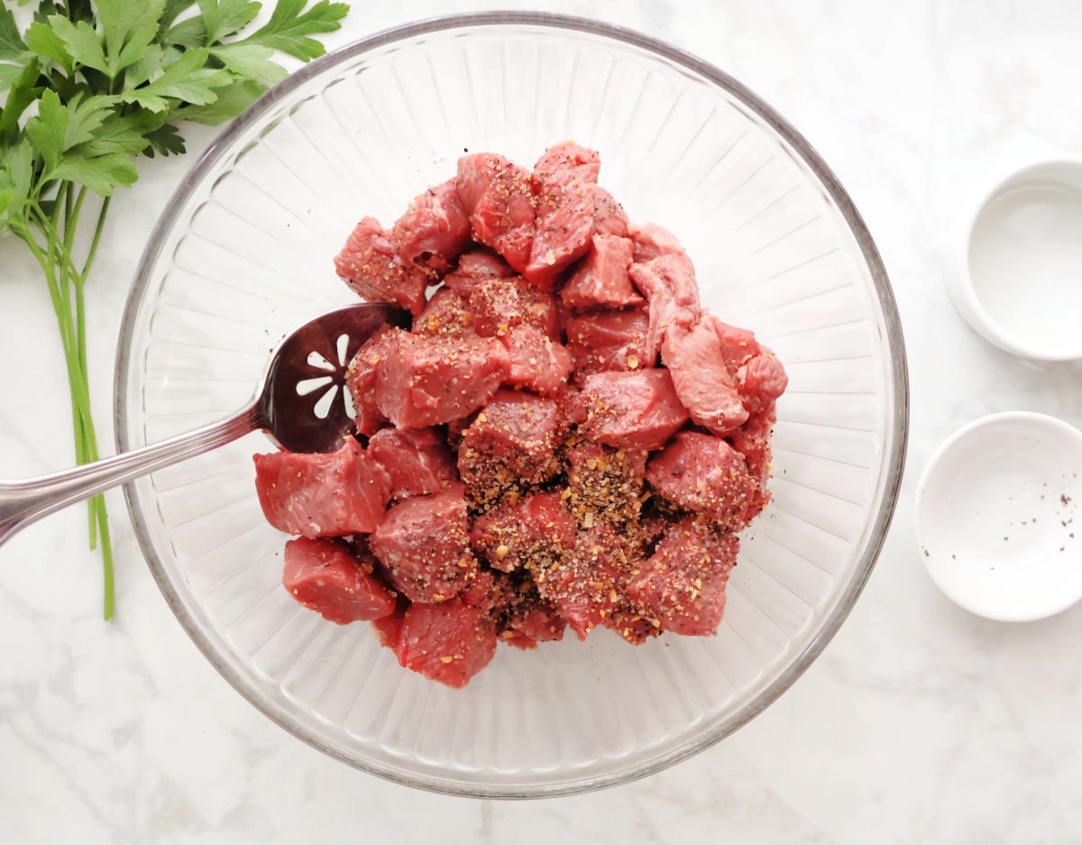 A bowl of bite sized pieces of uncooked steak gets olive oil and seasoning mixed into it. 