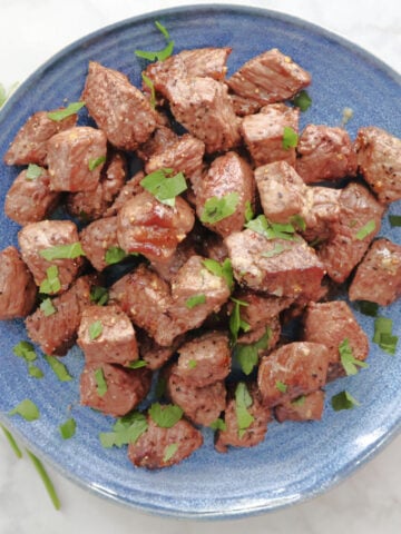 A plate of steak bites garnished with small pieces of parsley. The Montreal Steak Seasoning and the juices of the steak are visible on the blue serving plate.