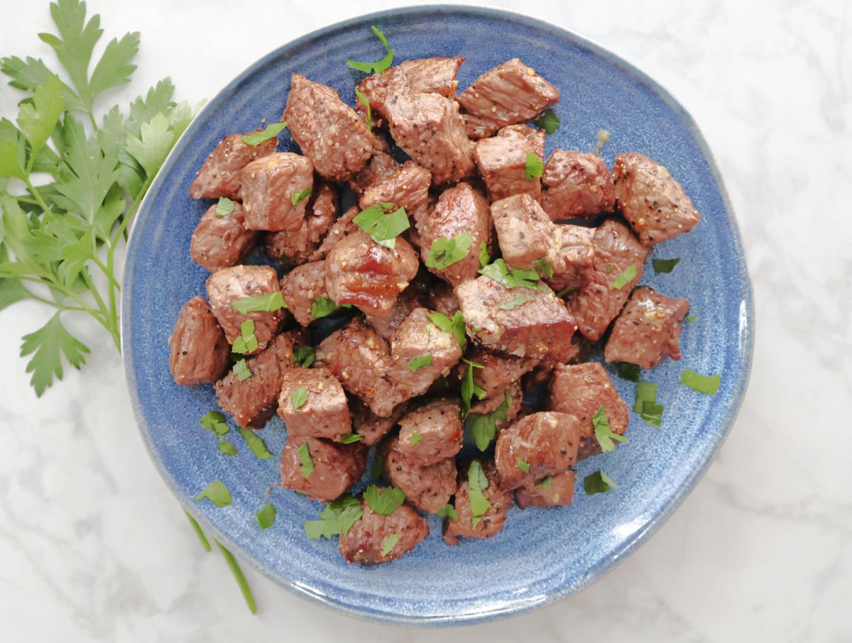 A plate of steak bites garnished with small pieces of parsley. The Montreal Steak Seasoning and the juices of the steak are visible on the blue serving plate.  