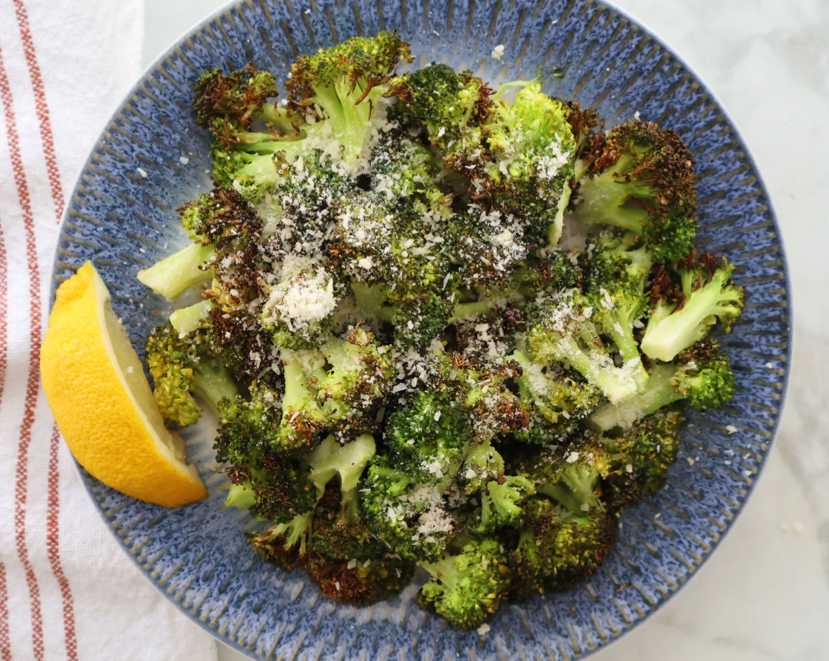 air fried broccoli with a sprinkling of parmesan cheese and a drizzle of lemon. Lemon wedge is to the side of the broccoli and it is all served on a blue plate.