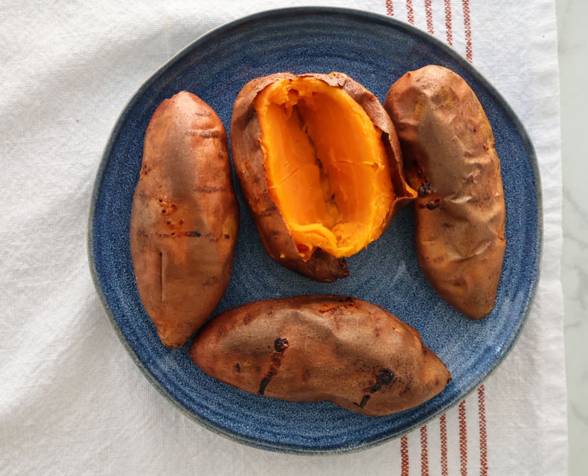 Four Sweet potatoes on a plate. One has been cut into so you can see the cooked orange interior.