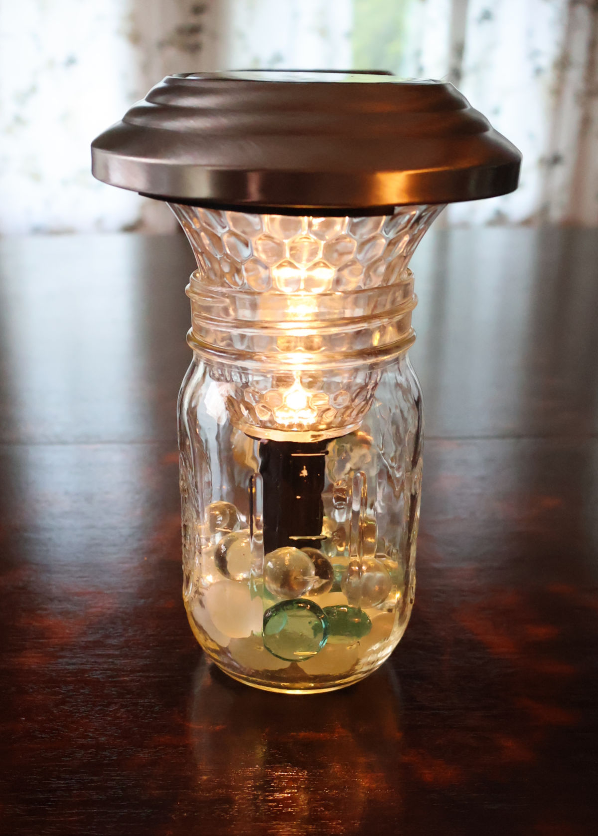 Mason jar lamp with mason jar with marbles and the top of an outdoor landscaping solar light. Light is glowing on a table.