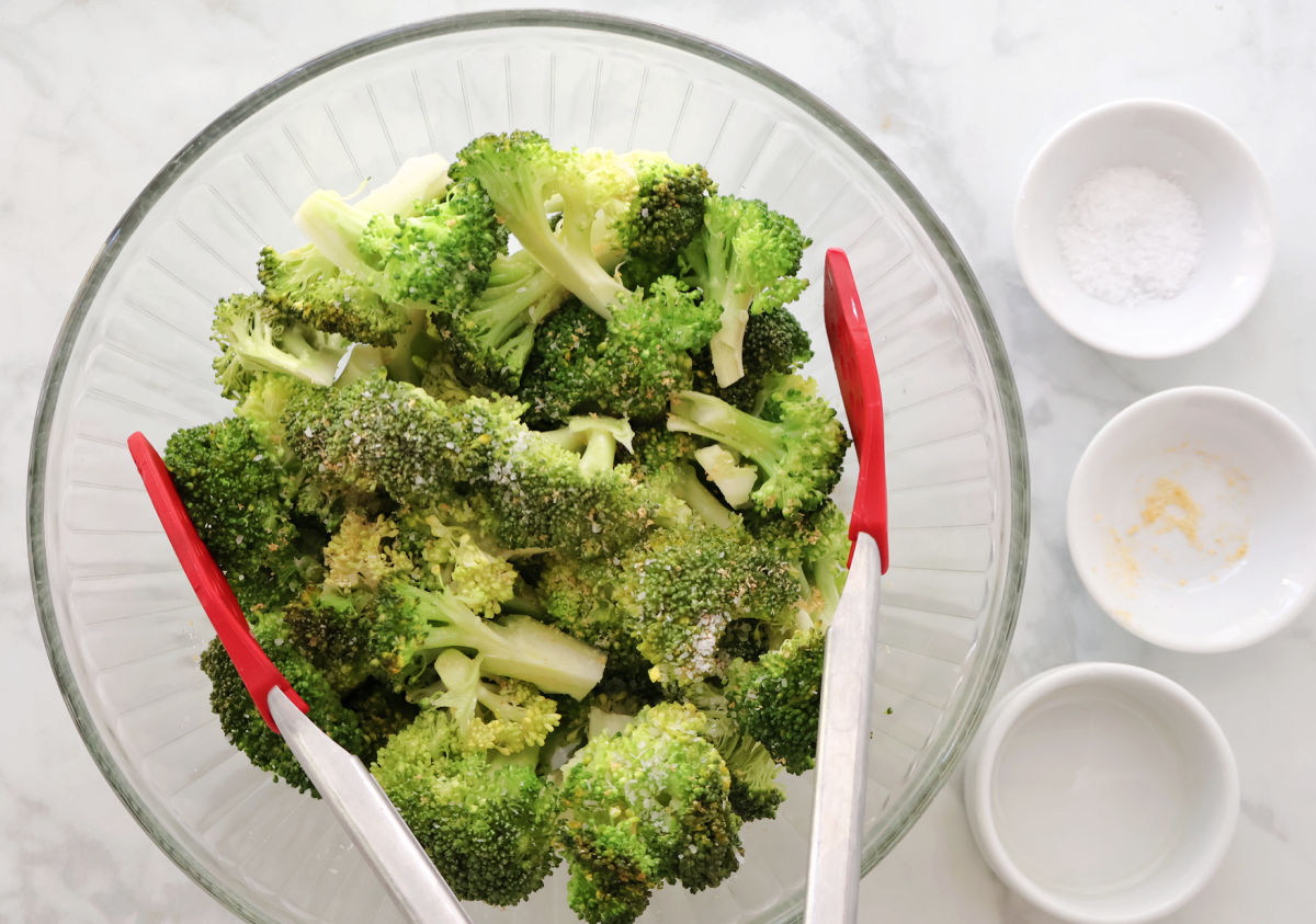 broccoli topped with garlic powder, Kosher salt and olive oil in a clear glass bowl. Red tipped tongs are used to mix. 