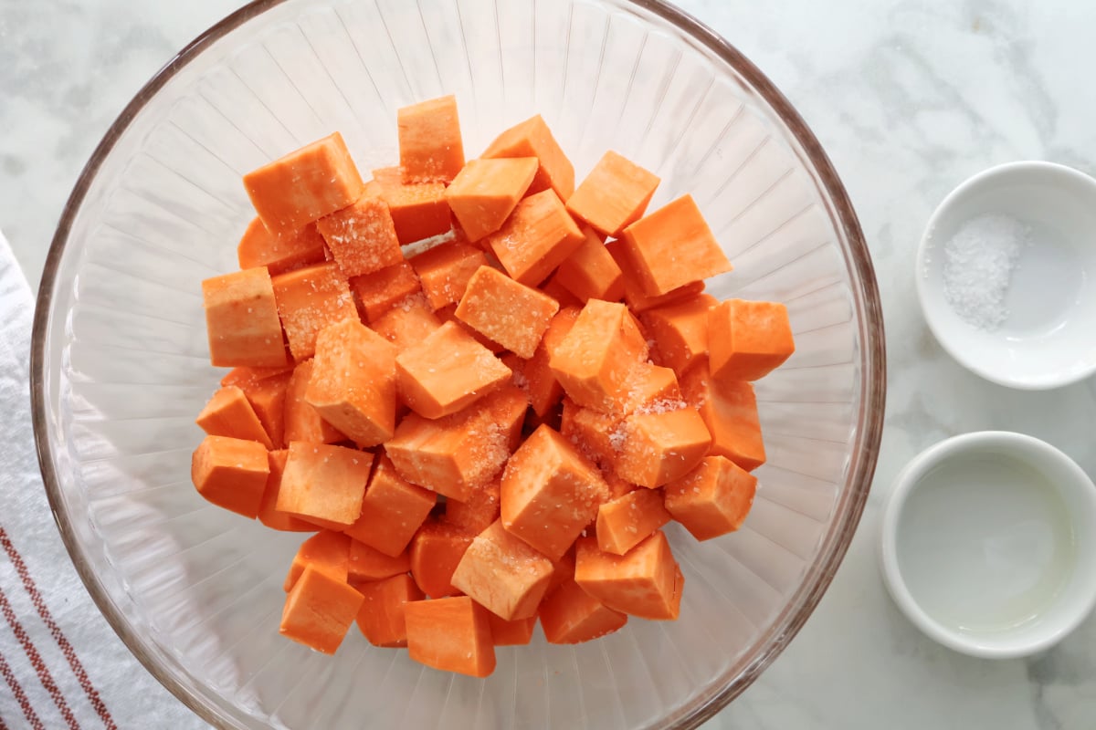 Cubed sweet potato that has olive oil poured on and kosher salt sprinkled on top. In a clear glass bowl.