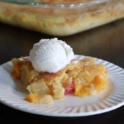 A portion of fruit cocktail dump cake on a white plate. The baking dish full of the fruit cocktail dump cake is visible behind the plate.