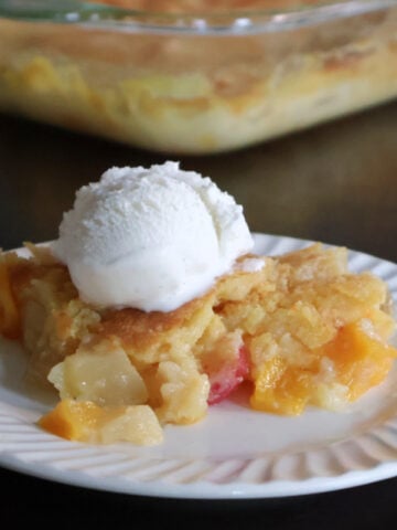 A portion of fruit cocktail dump cake on a white plate. The baking dish full of the fruit cocktail dump cake is visible behind the plate.