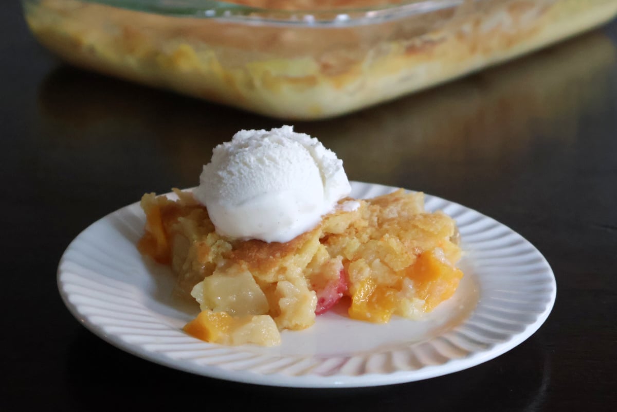 A portion of fruit cocktail dump cake on a white plate. The baking dish full of the fruit cocktail dump cake is visible behind the plate.