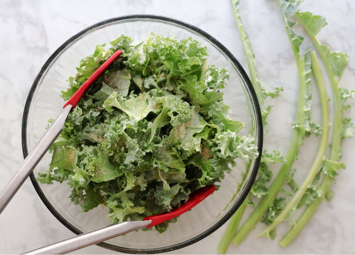 Torn kale for air fried kale in a glass bowl with seasonings ready to be mixed in with red tipped tongs.