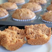 A split carrot cake muffin allowing you to see the pineapple chunks. Behind the muffin is the muffin tin full of carrot muffins.