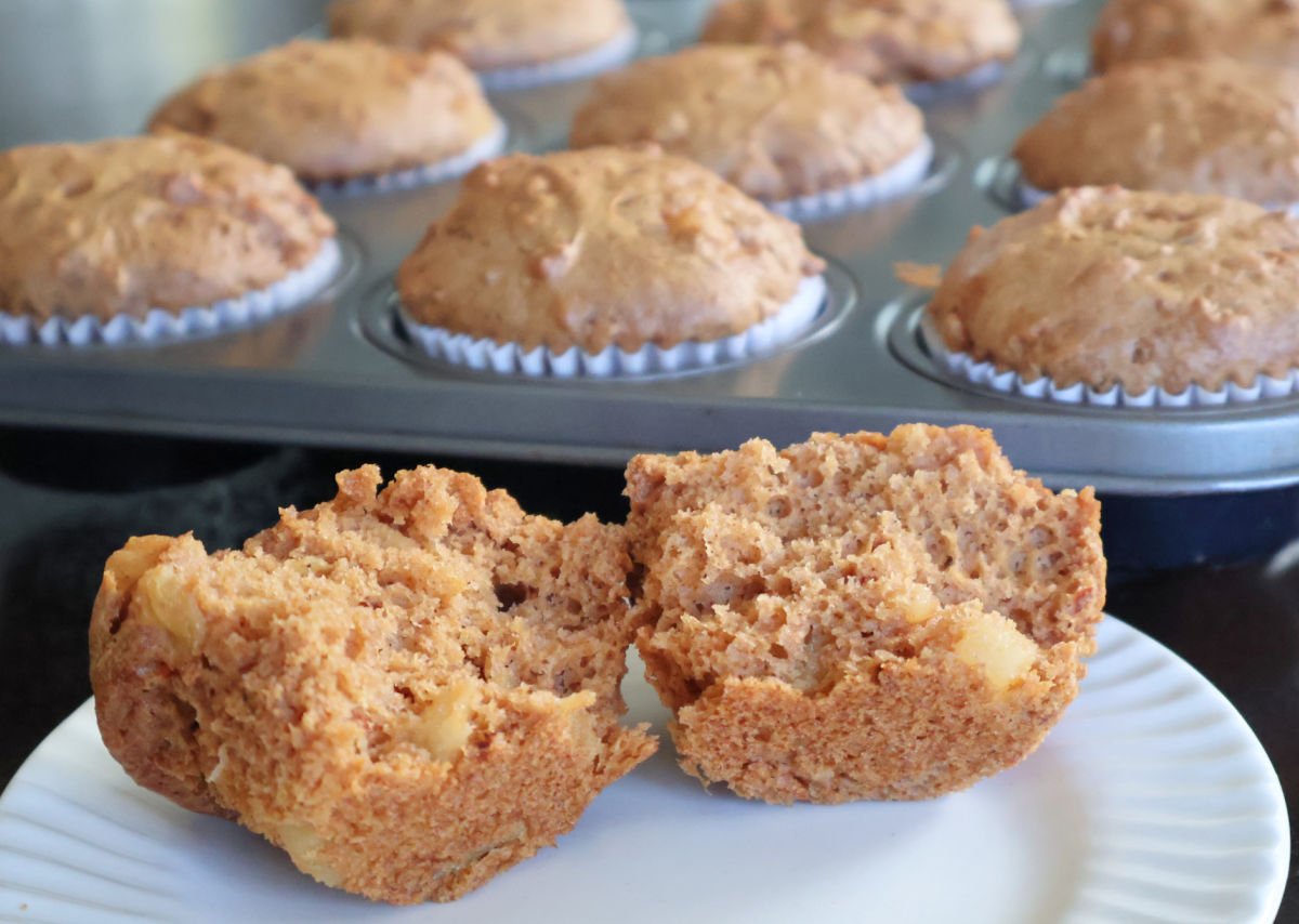 A split carrot cake muffin allowing you to see the pineapple chunks. Behind the muffin is the muffin tin full of carrot muffins. 