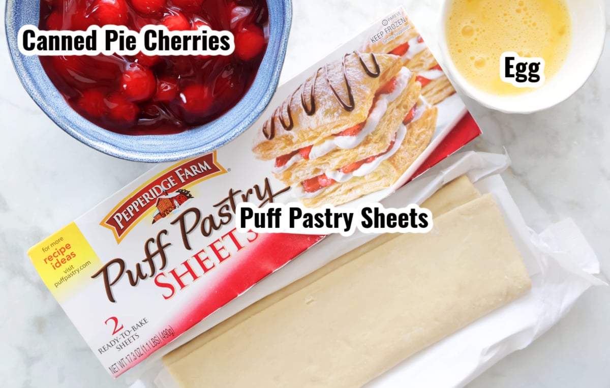 Two ingredients to make cherry strudel with puff pastry.  Image shows bowl of cherry pie filling in a blue bowl, the box of puff pastry sheets, with the pastry taken out of the box and a beaten egg for the egg wash. 