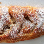 Apple strudel on a white tray. It has slits cut into the top and the apples are visible. Powdered sugar has been sprinkled on top.