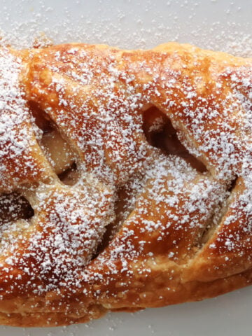 Apple strudel on a white tray. It has slits cut into the top and the apples are visible. Powdered sugar has been sprinkled on top.