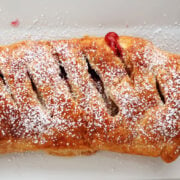 A cherry strudel made with puff pastry with cherry pie filling peeking through slits in the pastry. Cherry strudel has been dusted with icing sugar and is being served on a white tray.