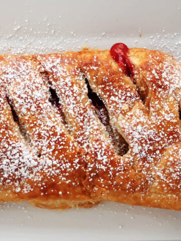 A cherry strudel made with puff pastry with cherry pie filling peeking through slits in the pastry. Cherry strudel has been dusted with icing sugar and is being served on a white tray.