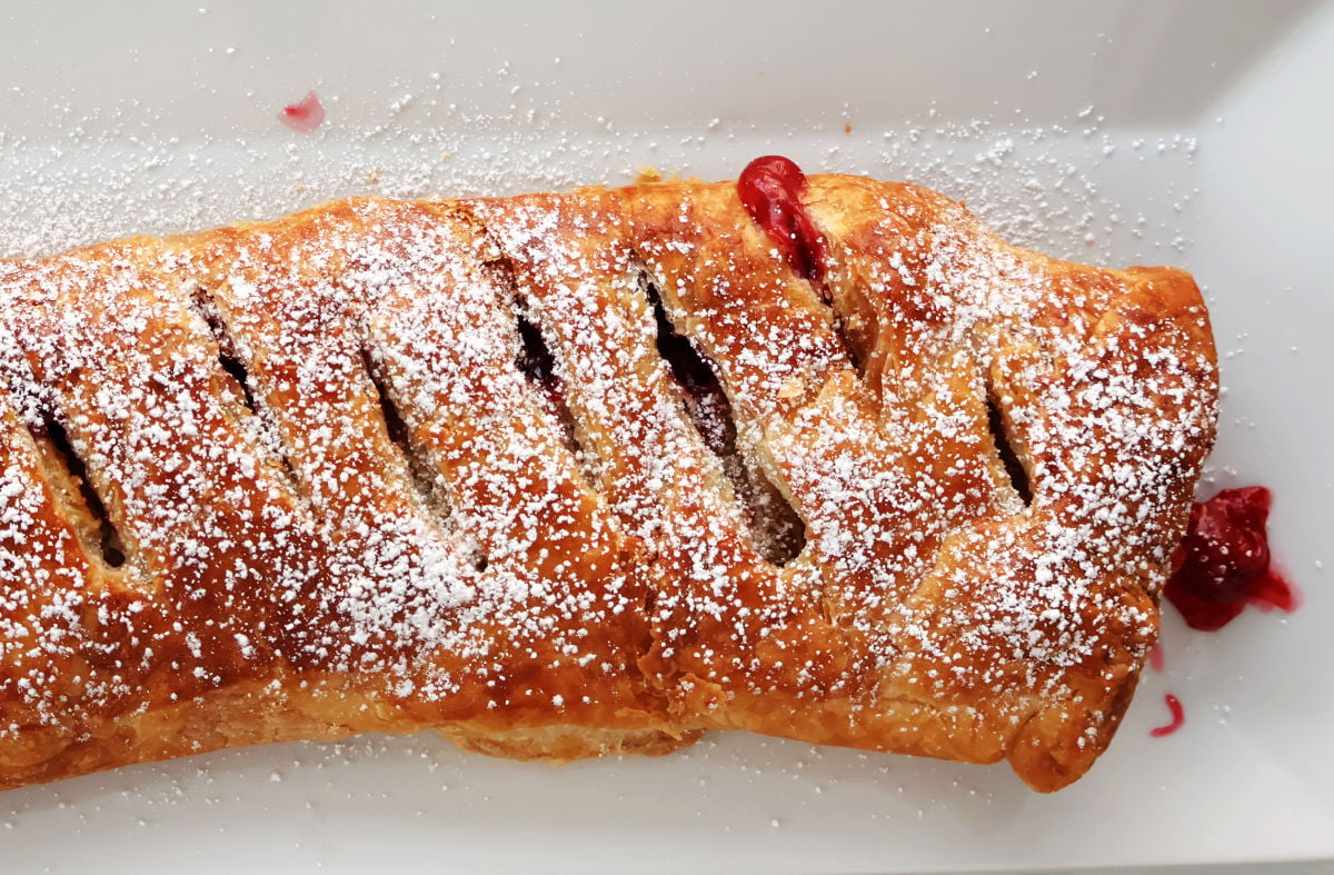 A cherry strudel made with puff pastry with cherry pie filling peeking through slits in the pastry. Cherry strudel has been dusted with icing sugar and is being served on a white tray.