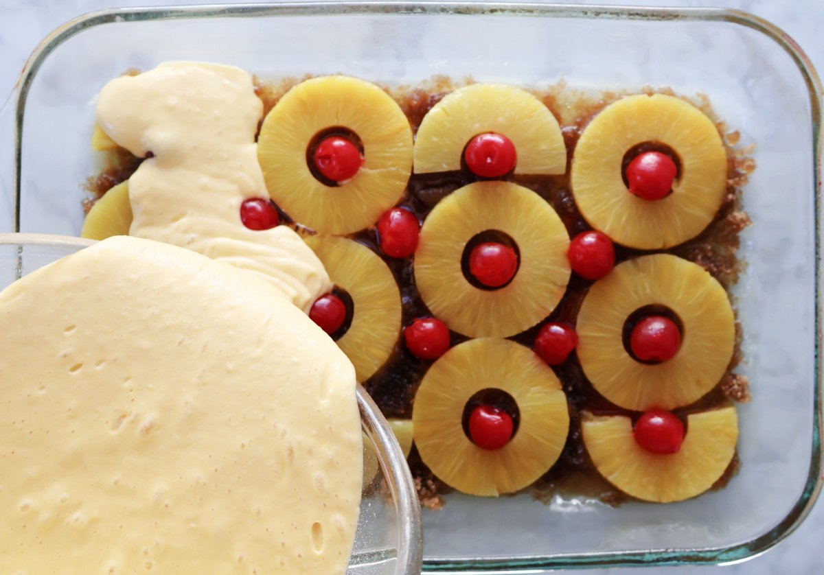 Pouring cake batter on top of pineapple slices and cherries to assemble the pineapple upside down cake.