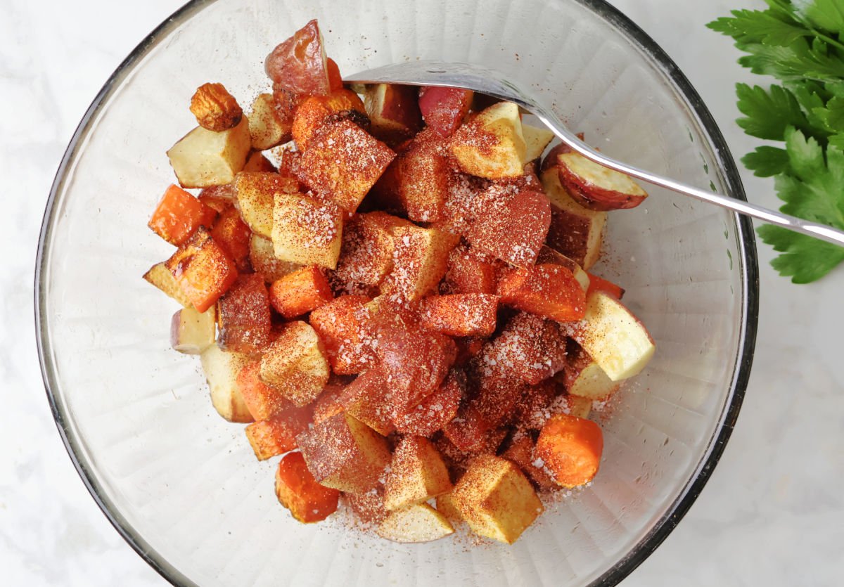 Adding seasonings including spiced paprika to cubed red potatoes and carrots. 
Spoon is in the bowl ready to mix seasonings onto veggies.