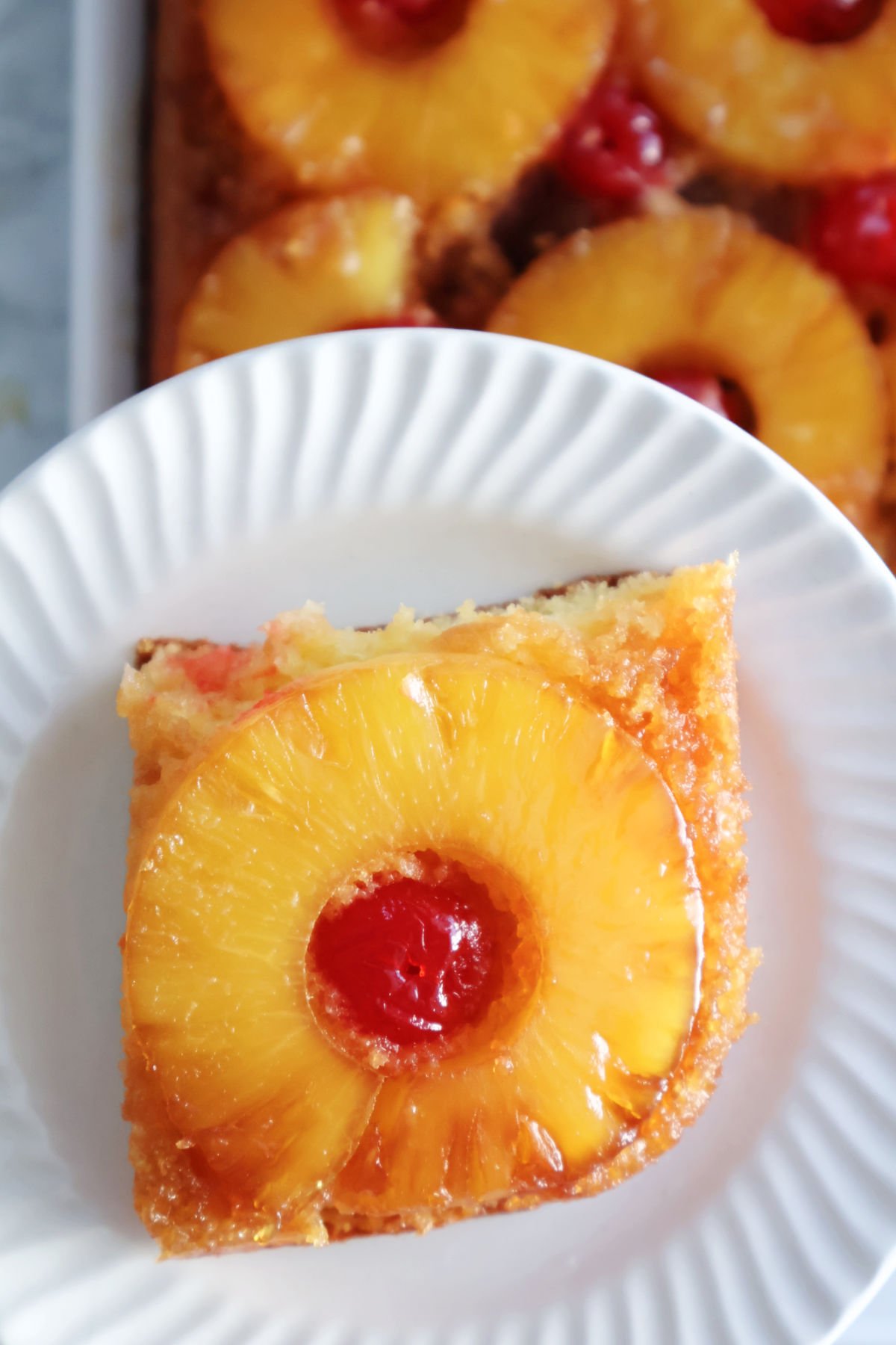 A serving of pineapple upside down cake showing a ring of pineapple with a maraschino cherry in the center of the pineapple ring. The delicious caramel topping created by the first layer of brown sugar and butter is visible on top of the cake and running down the sides.