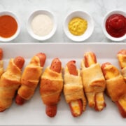 Hot dogs rolled in crescent roll dough baked to a golden brown. Served on a white tray with ketchup, mustard, ranch dressing and bbq sauce for dipping.