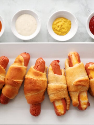 Hot dogs rolled in crescent roll dough baked to a golden brown. Served on a white tray with ketchup, mustard, ranch dressing and bbq sauce for dipping.