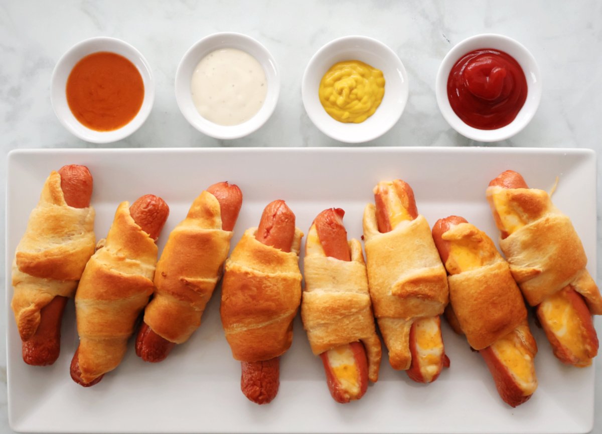 Hot dogs rolled in crescent roll dough baked to a golden brown. Served on a white tray with ketchup, mustard, ranch dressing and bbq sauce for dipping.