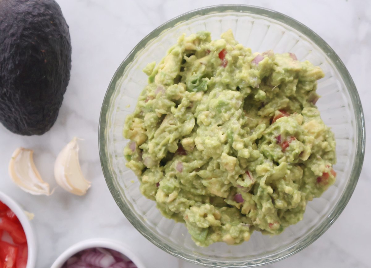 fresh guacamole in a clear glass bowl surrounded by fresh ingredients including avocado, chopped tomato, chopped onion and garlic